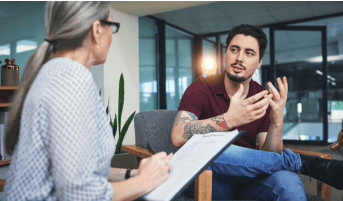 Man with tattooed arm seated and speaking to a woman with a clipboard