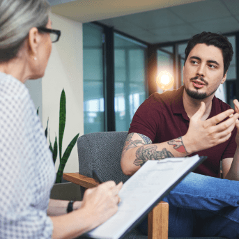 Man with tattooed arm seated and speaking to a woman with a clipboard